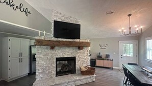 A modern kitchen with a stone fireplace, white cabinets, and a dining area with a wooden table and chairs. - Fireplace Lodge - 1 mile from Golf Course & 5 mins from the lake! (Isabella)