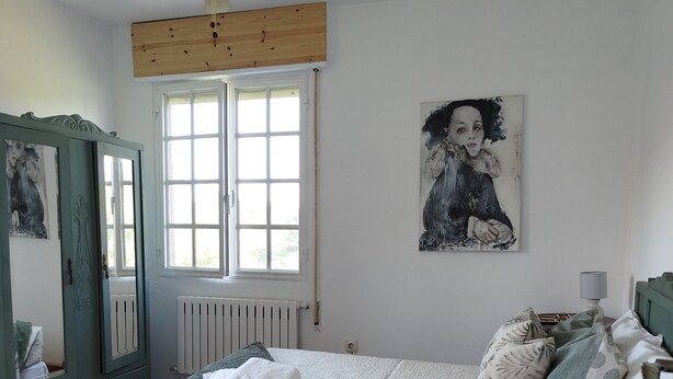 Un dormitorio con cama, ventilador de techo, ventana, radiador, cómoda y un cuadro en la pared.