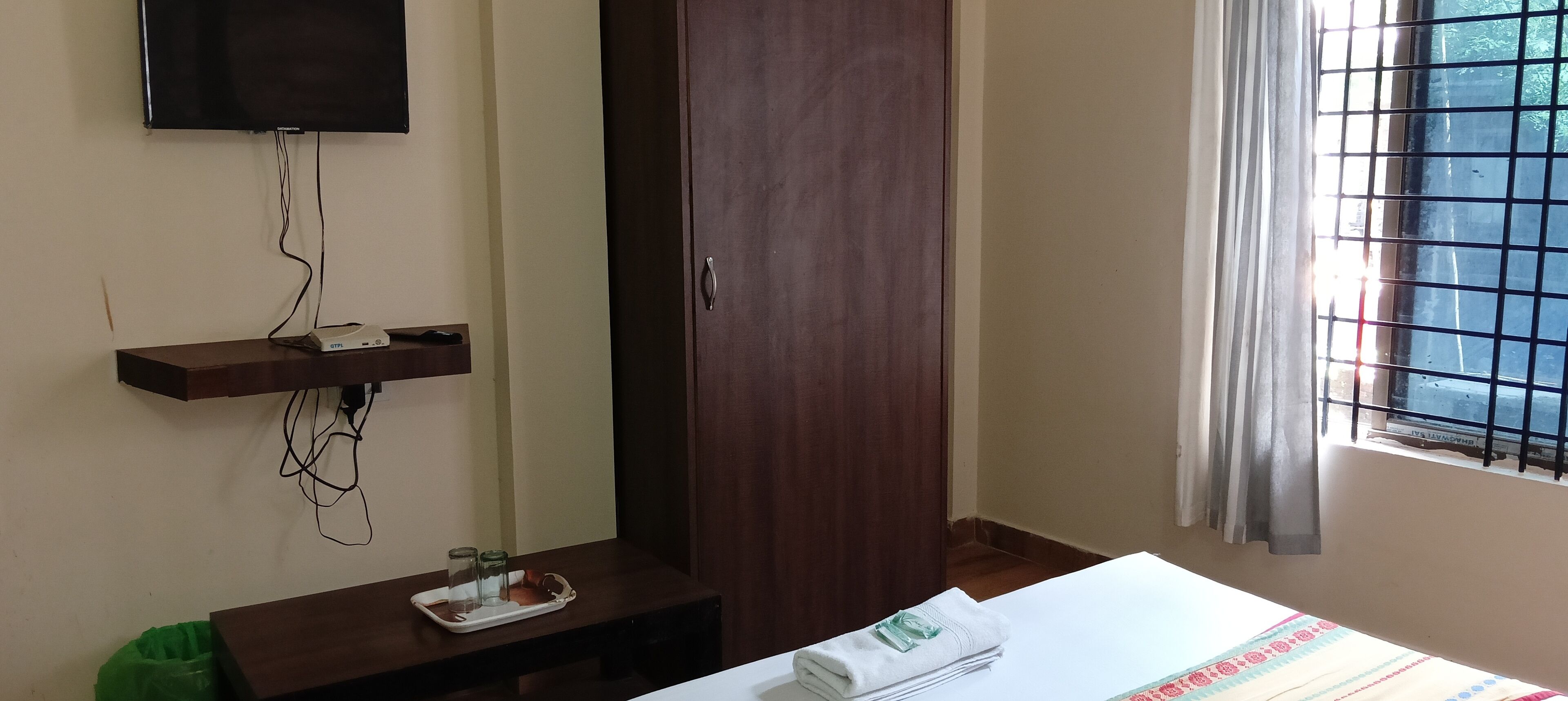 Habitació amb un armari de fusta, televisió penjada a la paret, finestra amb reixeta metàl·lica i llit amb una tovallola blanca arremolinada amb cura.