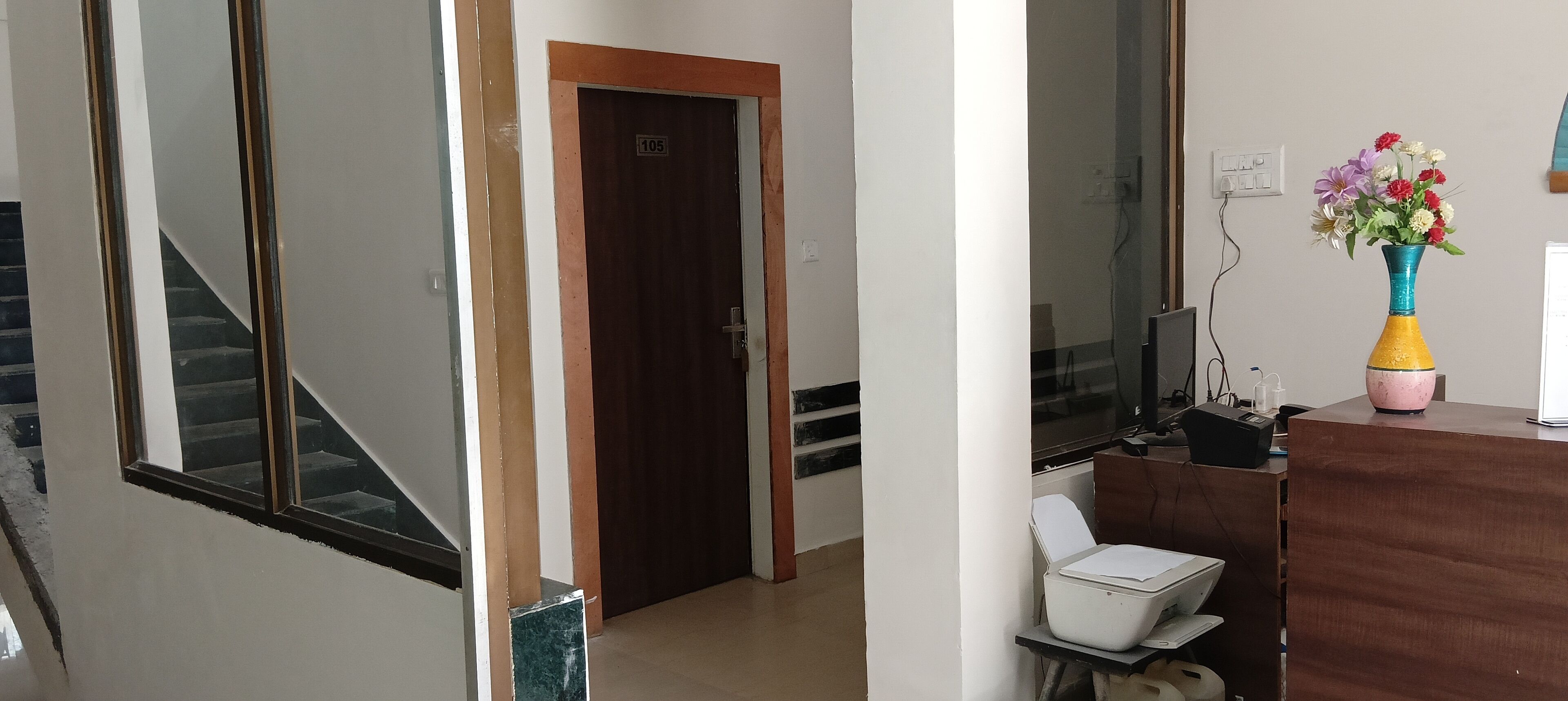 A hotel room with a wooden door, a desk with a printer, a chair, and a vase of flowers.