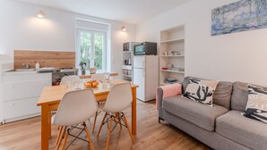 Una cocina moderna con encimera de madera, armarios blancos y un comedor con mesa y sillas de madera. En la sala de estar, un sofá gris con cojines estampados.