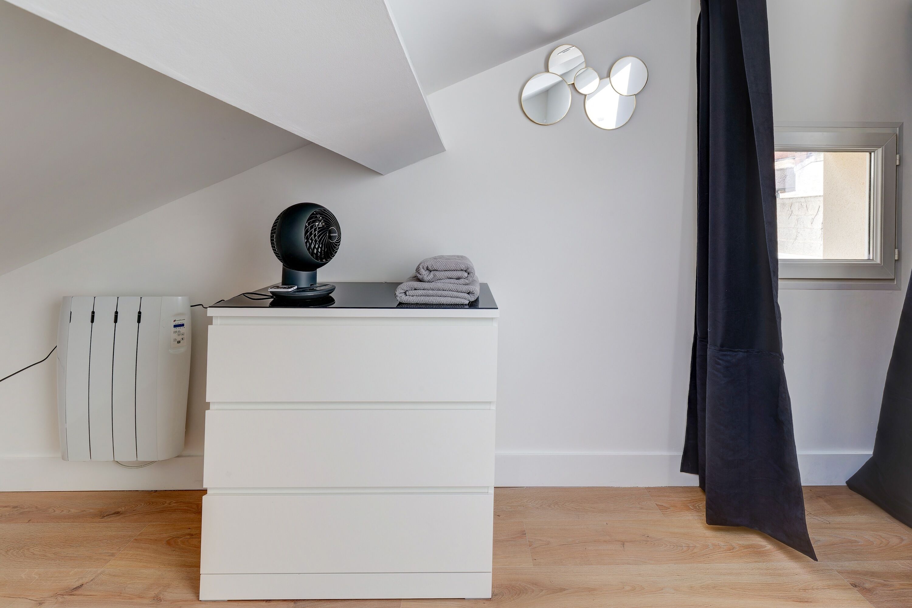 A modern room with a white chest of drawers, a black hairdryer, and a window with a black curtain.