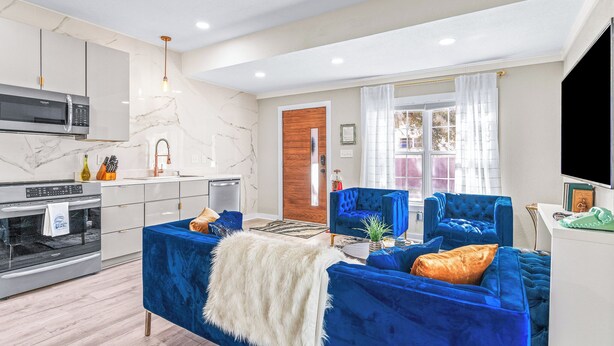 Una sala de estar moderna con un sofá azul, un manto de piel blanca y una cocina con armarios blancos y un salpicadero de mármol.