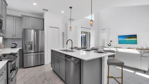 A modern kitchen with stainless steel appliances, a central island with a sink, and a dining area with a white sofa and a TV displaying a tropical scene. - Batman | Mickey | Game Room | 4 Miles Disney 2669 (Kissimmee)