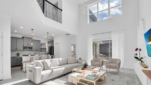 A spacious living area with a white sofa, a coffee table, and a dining table. There is a kitchen in the background with modern appliances. - Batman | Mickey | Game Room | 4 Miles Disney 2669 (Kissimmee)
