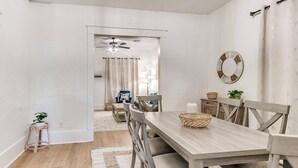 A dining area with a wooden table and chairs, a ceiling fan, and a view into another room with a ceiling fan and curtains.