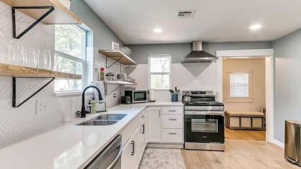 A modern kitchen with white cabinetry, a stainless steel stove, and a microwave. There are wooden shelves above the counter and a window with a view of trees.