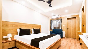 A modern bedroom with a wooden bed, a ceiling fan, an air conditioning unit, a blue chair, and a white bedside table with a lamp. - Townhouse Shree Jagannath Puri (Puri)