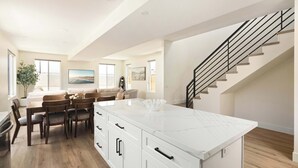 A spacious kitchen with a large island, wooden dining table, and chairs, a staircase, and a view of the outdoors. - Coastal Ocean Beach Compound Escape (San Diego)
