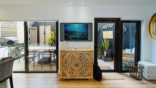 Een moderne woonkamer met een glazen schuifpui naar een balkon, een houten kast met gedetailleerde houtsnijwerken, een flatscreen televisie aan de wand en een eethoek met tafel en stoelen.