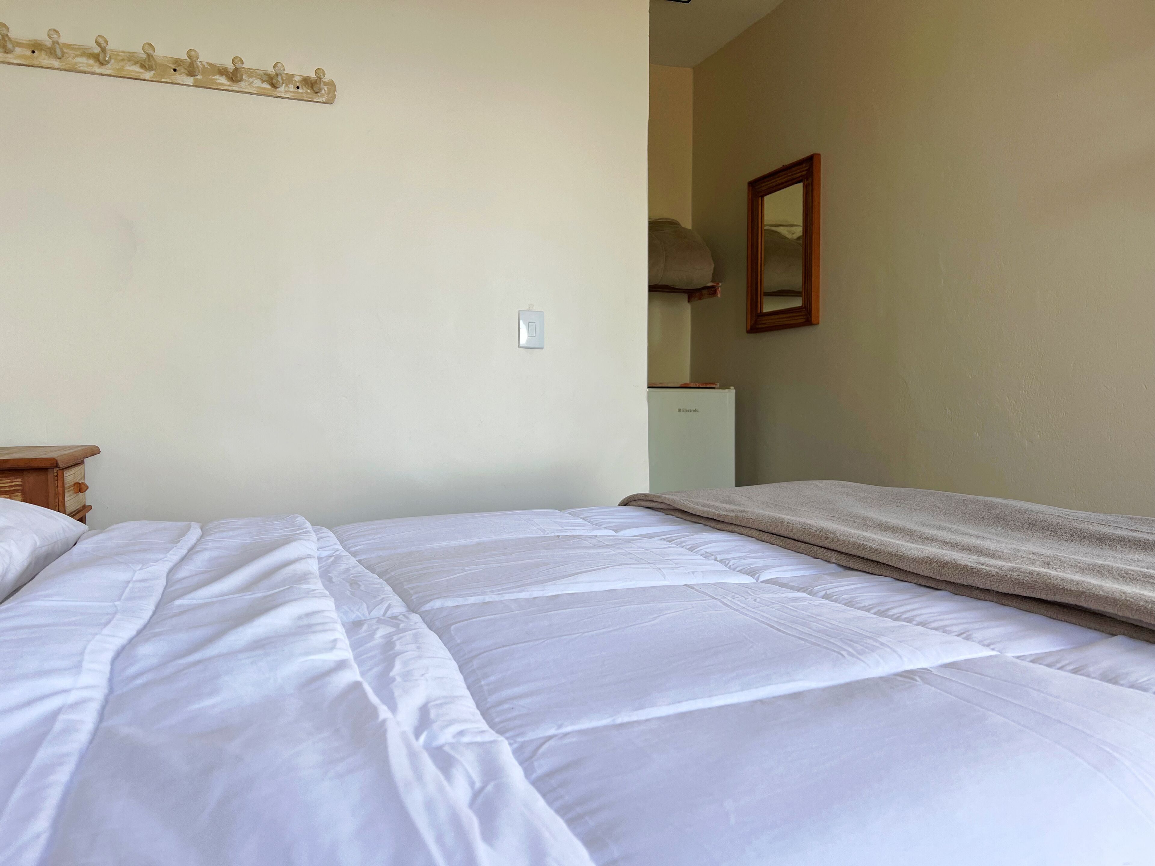 A neatly made bed with white bedding, a wooden nightstand, a small table, and a wall-mounted light switch.