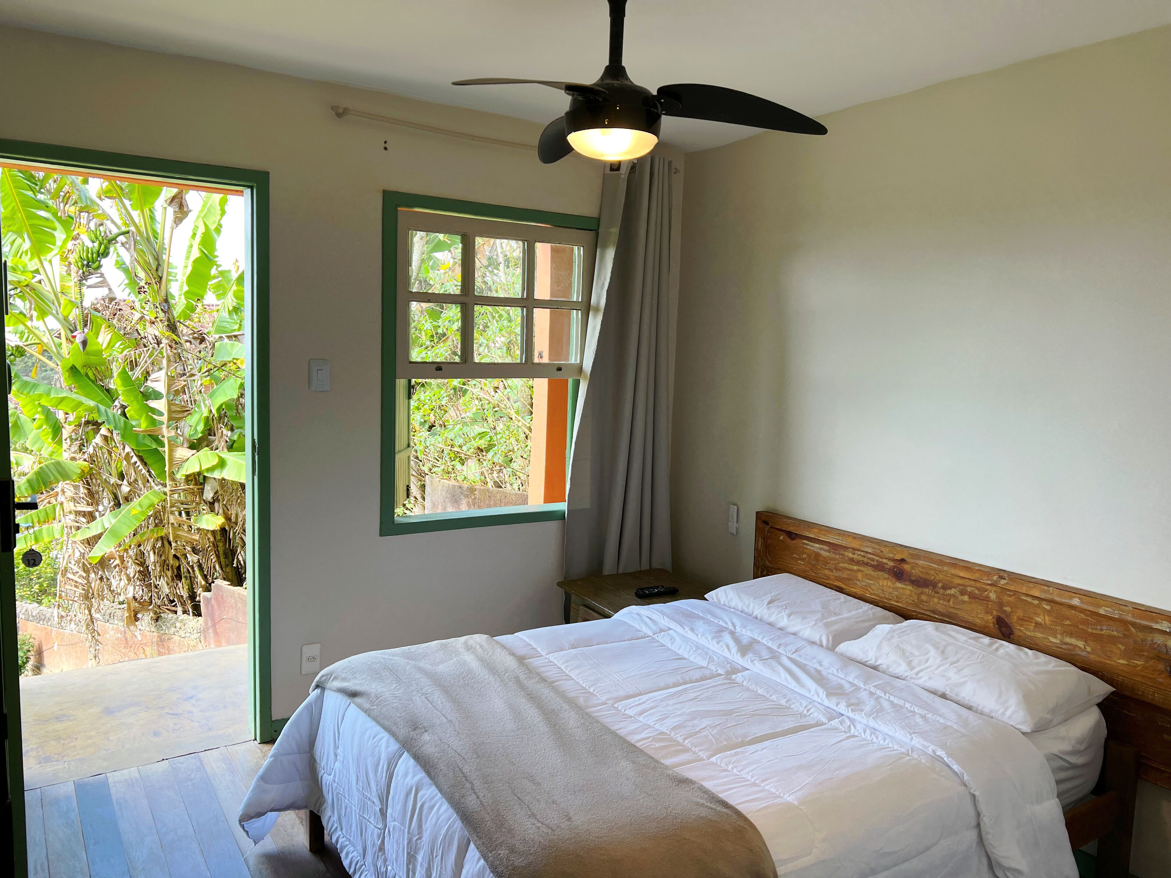 Un dormitorio con cama, ventilador de techo, ventana con cortinas y vista a follaje exterior.