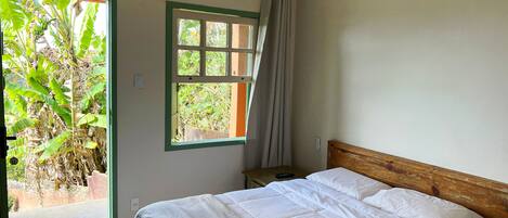 Un dormitorio con cama, ventilador de techo, ventana con cortinas y vista a follaje exterior.