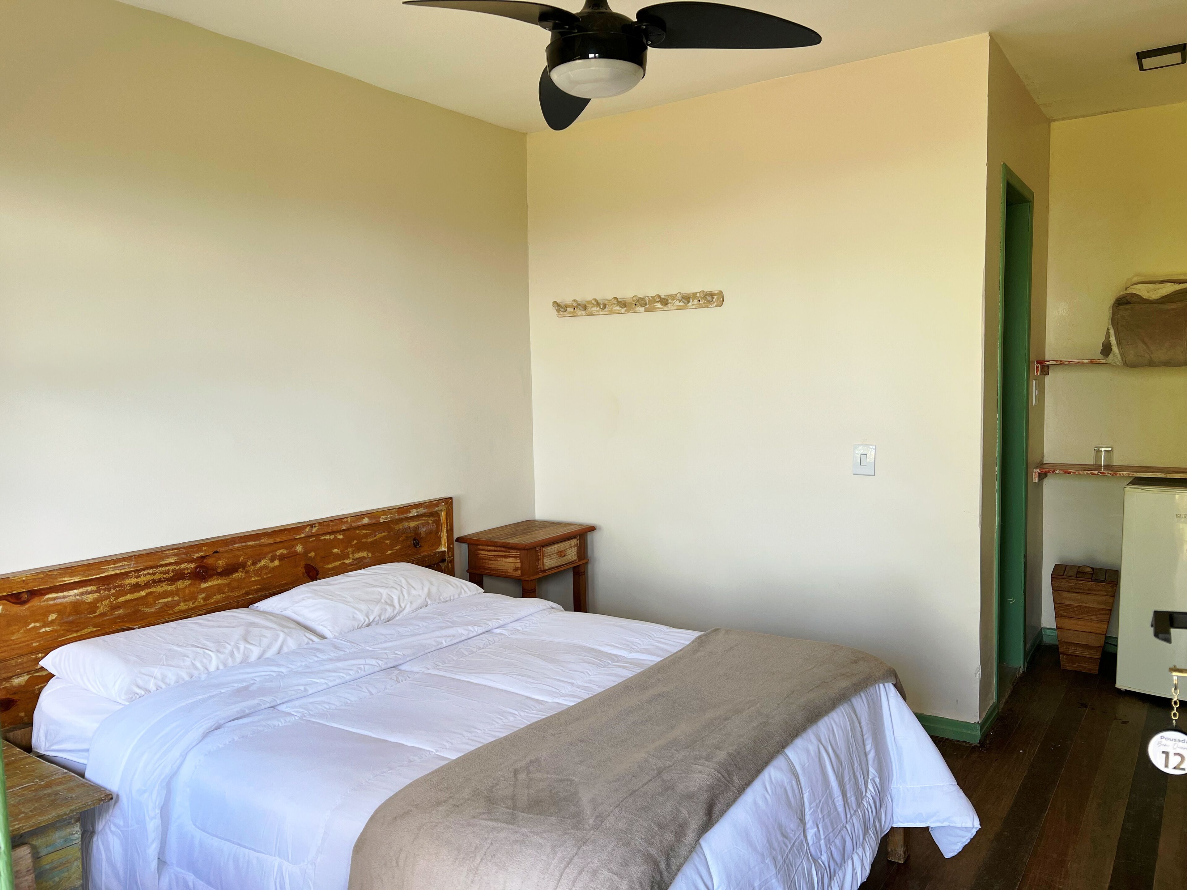 Una camera da letto con una testiera in legno, un letto singolo con biancheria bianca, un comodino in legno e un ventilatore a soffitto, e una porta verde che dà su un'altra stanza.