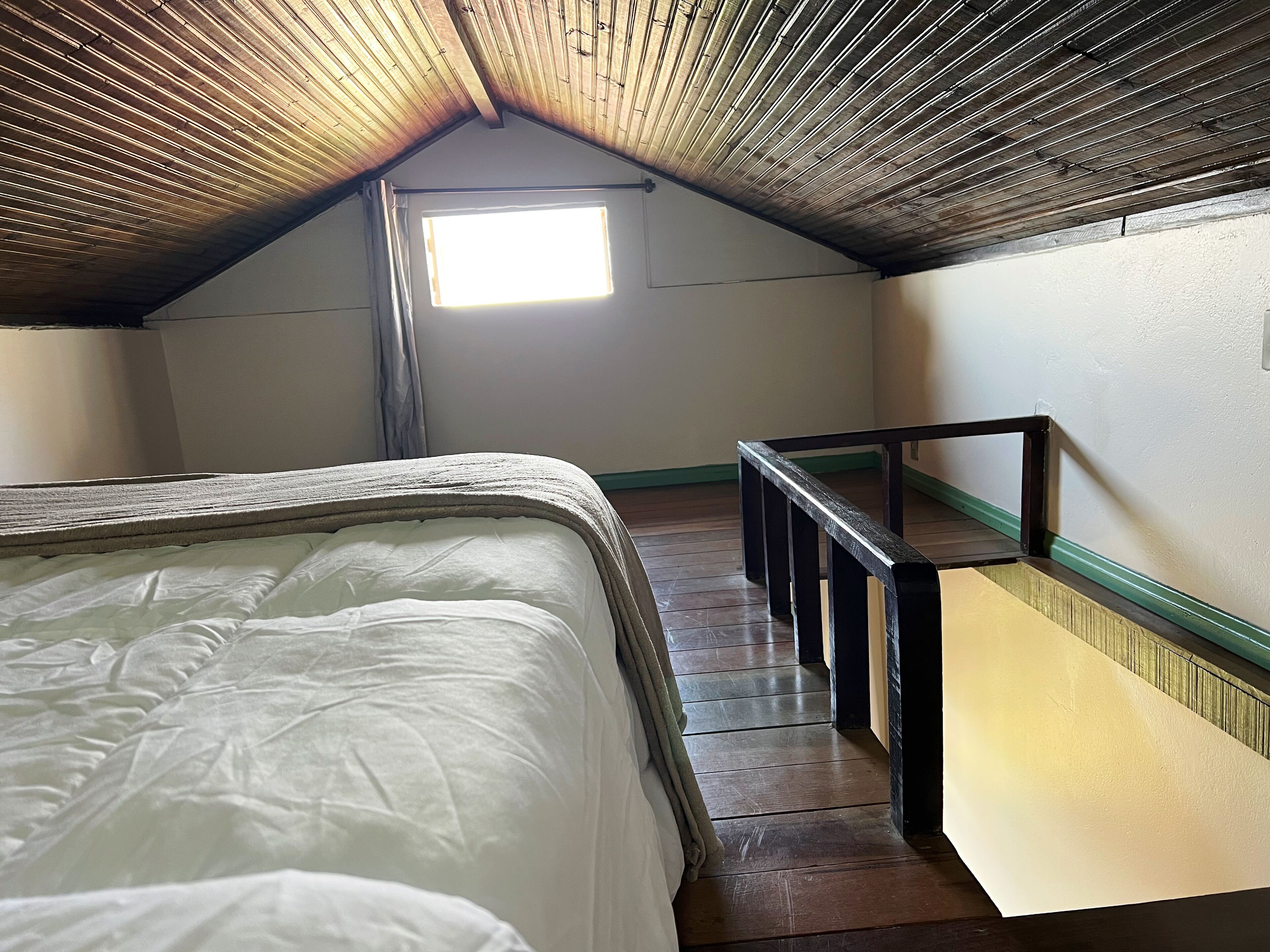 Un dormitorio bajo los tejados, estrecho, con techo de madera, una cama individual, un ventanal pequeño y una barandilla de madera.