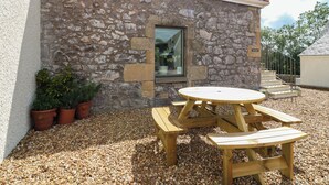 A stone building with a tiled roof, a window, and a wooden table and bench set on a gravel surface.