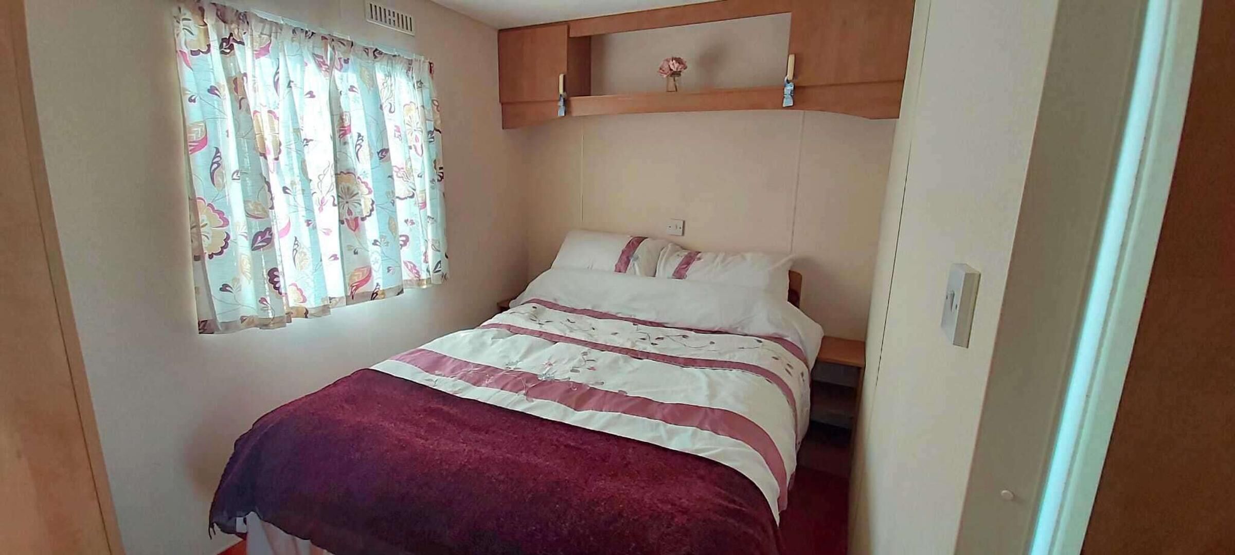 A bedroom with a bed, a window with floral curtains, and a wooden shelf above the bed.