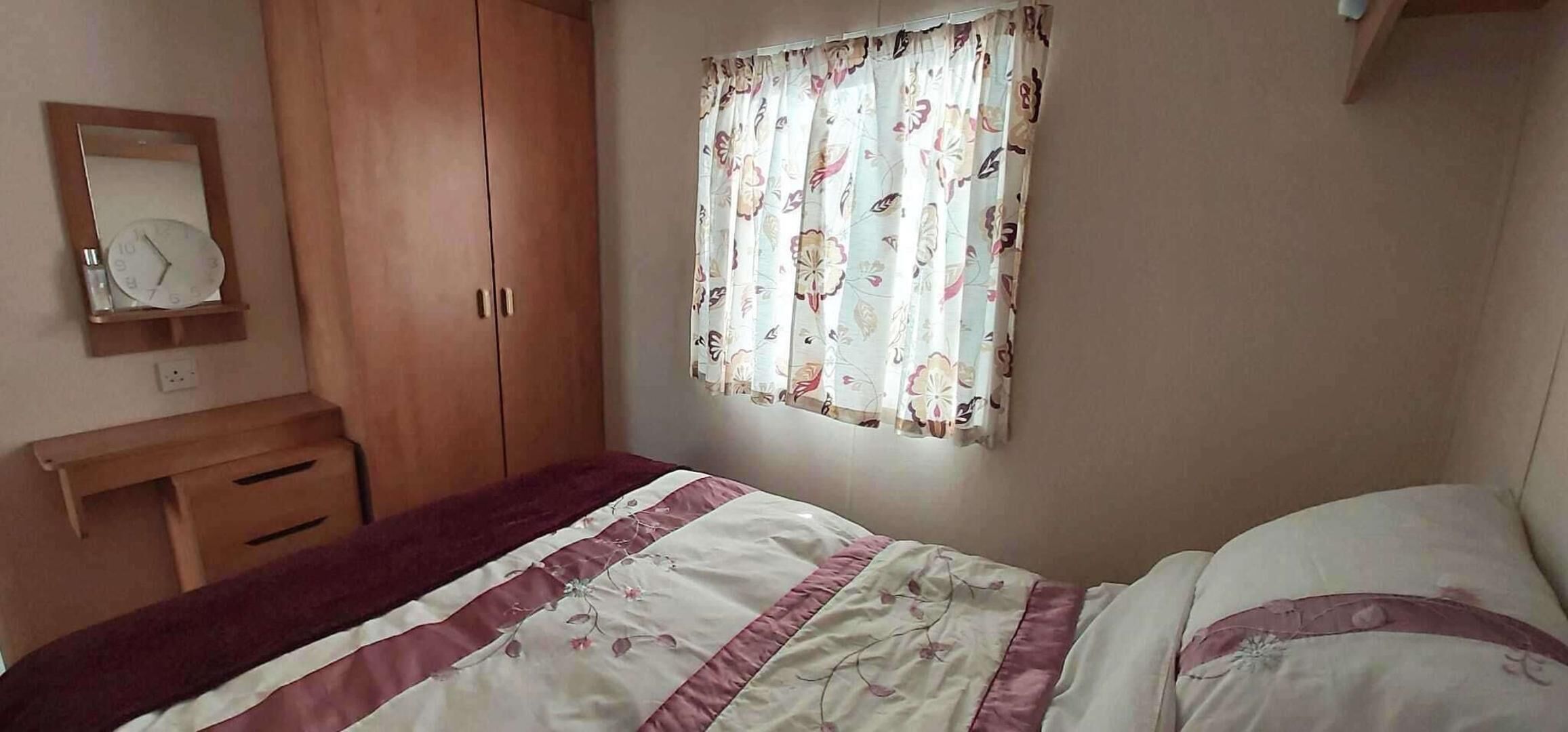 A bedroom with a bed, a wardrobe, a mirror, and a window with floral curtains.