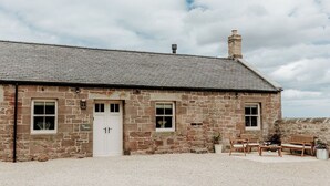 A stone building with a white door and windows, a gravel driveway, and outdoor seating. - Step into thoughtfully restored accommodation a stone's throw from Holy Island (Beal)