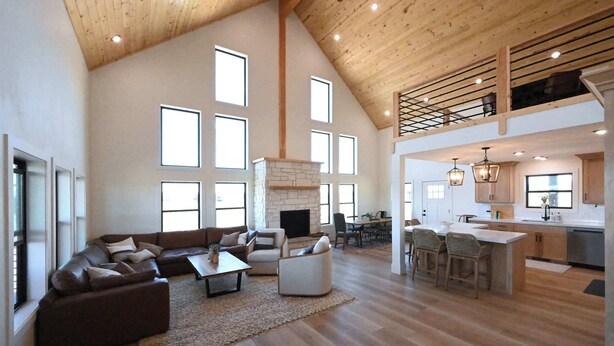A spacious living area with a high ceiling, large windows, a fireplace, and a kitchen with bar stools.