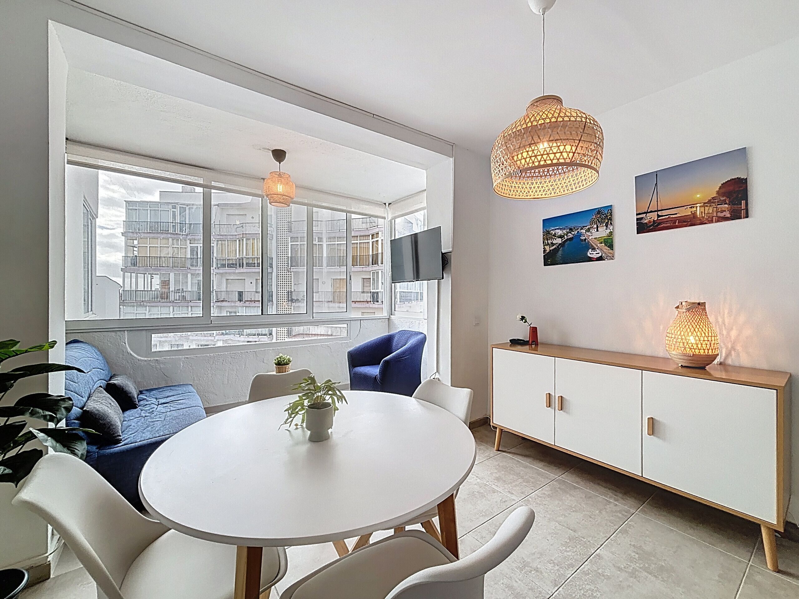 Una sala de estar moderna con una mesa de comedor redonda, sillas azules, un armario blanco y un ventanal con vistas a la ciudad.