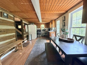 A cabin interior with a wooden ceiling, a dining table with chairs, a kitchen area with appliances, and a window with a view of trees.
