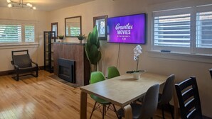 Ein modernes Wohnzimmer mit Holzboden, Kamin, Esstisch mit Stühlen, Wand-TV und großen Fenstern mit Jalousien.