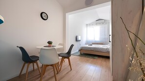 A modern bedroom with a bed, a bedside table, and a window. There is a dining area with a table and chairs. - Central City Apartments – Walk to Everything! (Podgorica)