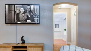 A modern living room with a flat-screen TV mounted on the wall, a wooden entertainment unit with a remote control and a decorative item, and a hallway leading to another room. - Charming & Cozy 3-bedroom house in wonderful Springfield with AC (Springfield)