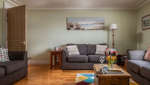 Ein Wohnzimmer mit grauem Sofa, einem Couchtisch mit Büchern und Blumen, einem Gemälde mit Strandmotiv und einer Lampe.
