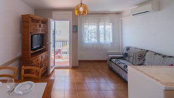 Een woonkamer met een televisie, een houten kast, een bank, een eettafel met stoelen, en een balkon met een hekwerk.