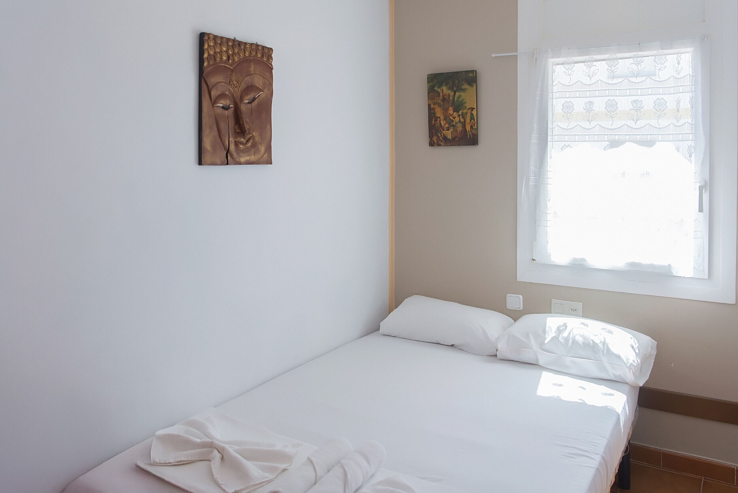 Una cama individual con ropa blanca, una ventana con cortinas translúcidas y dos obras de arte colgadas en la pared.
