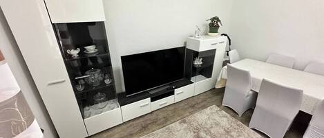 A modern living room with a flat-screen TV, a glass-fronted cabinet with decorative items, a dining area with a table and chairs, and a patterned rug on the floor.