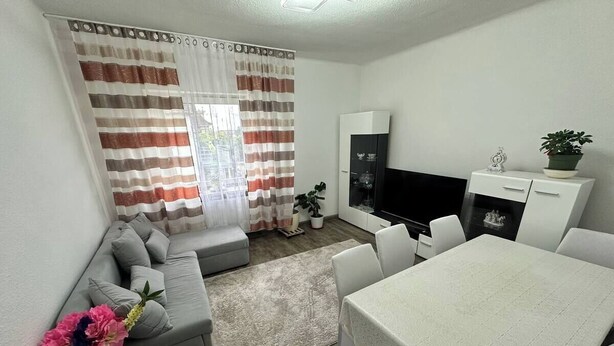 A modern living room with a grey sofa, a dining table with white chairs, a flat-screen TV, and a potted plant.