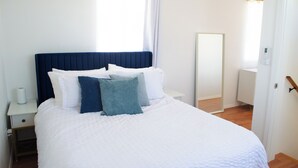A neatly made bed with white and blue bedding, a bedside table with a lamp, and a window with curtains.