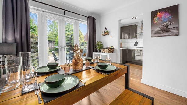 Un moderno comedor con una mesa de madera preparada para cuatro, una cocina al fondo y un ventanal con cortinas.