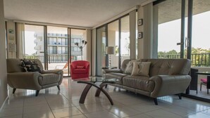 A modern living room with two sofas, a red chair, and a glass coffee table. Large windows offer views of a building and greenery.