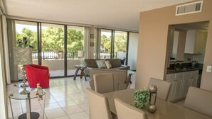 A spacious living area with a sofa, a red chair, and a glass coffee table. There is a balcony with a view of trees and a kitchen area visible through an open door.