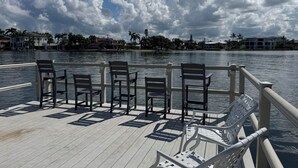 A waterfront deck with chairs and tables, overlooking a calm body of water with a cityscape in the background. - Mins to Beach, Tennis, Heated Pool, Bayside (Siesta Key)