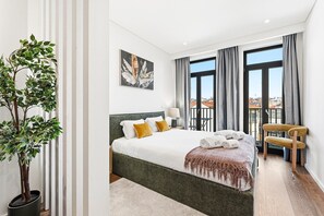 Habitación de hotel con cama, una planta en maceta, balcón con silla y vista a la ciudad.