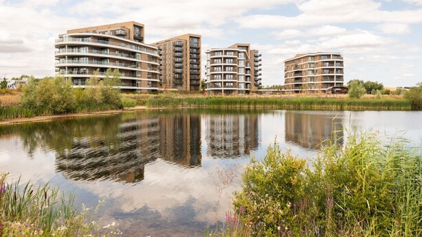 A modern residential complex with multiple high-rise buildings, a calm water body in the foreground, and a clear sky. - Modern 4BR New Home near Biz Park & Station | Pass the Keys (Reading)