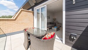 A patio with wicker furniture and a glass table, a sliding glass door leading to an interior living area with sofas and a lamp. - Modern 4BR New Home near Biz Park & Station | Pass the Keys (Reading)