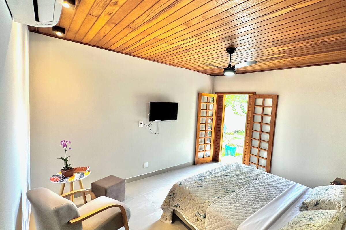 Een slaapkamer met een houten plafond, een bed met een gewatteerde spreideken, een stoel, een klein tafeltje met een plant en een aan de muur bevestigde televisie.