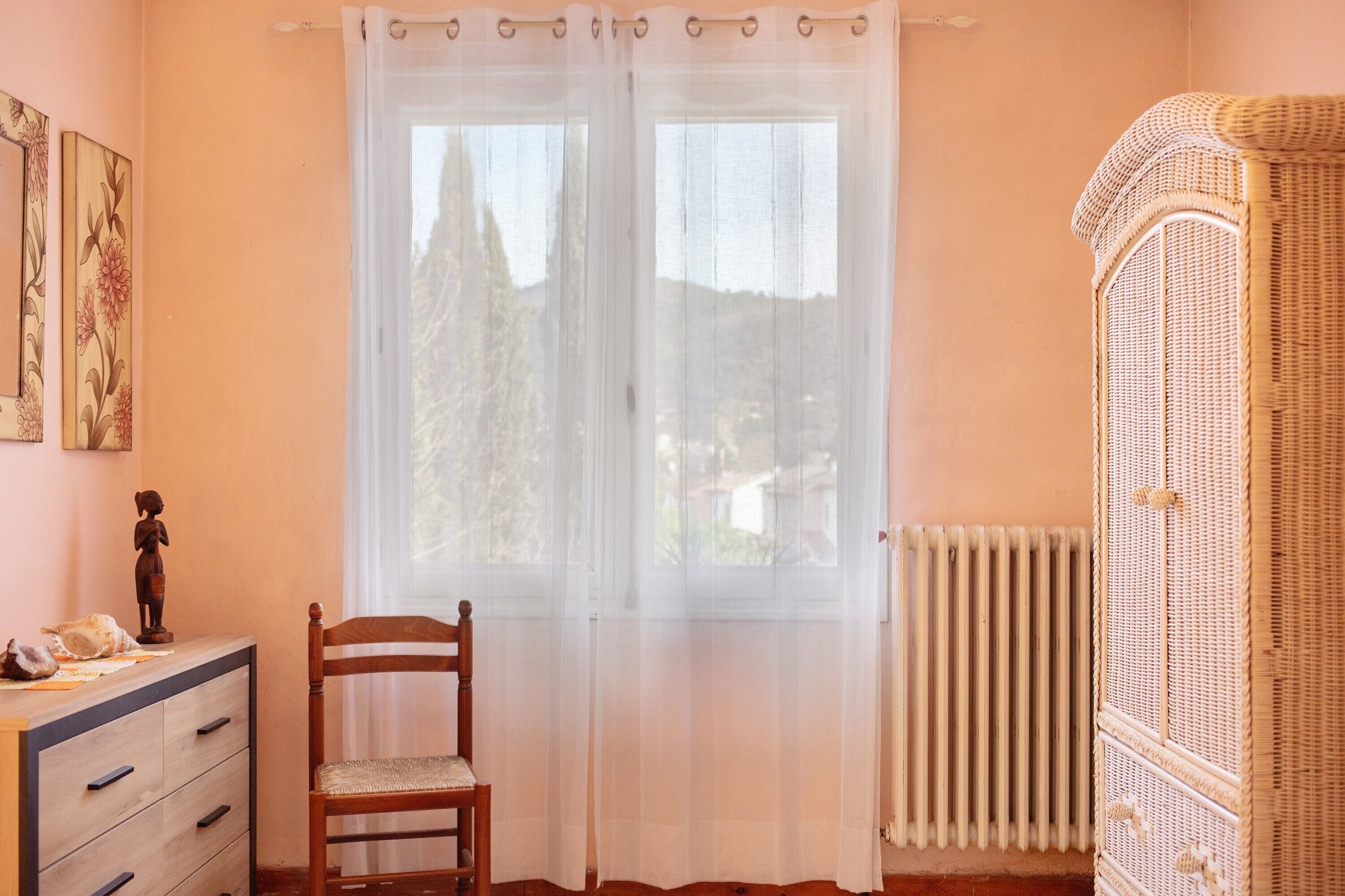 Une pièce comprenant une chaise en bois, une commode, un meuble en osier et une fenêtre ornée de rideaux légers.