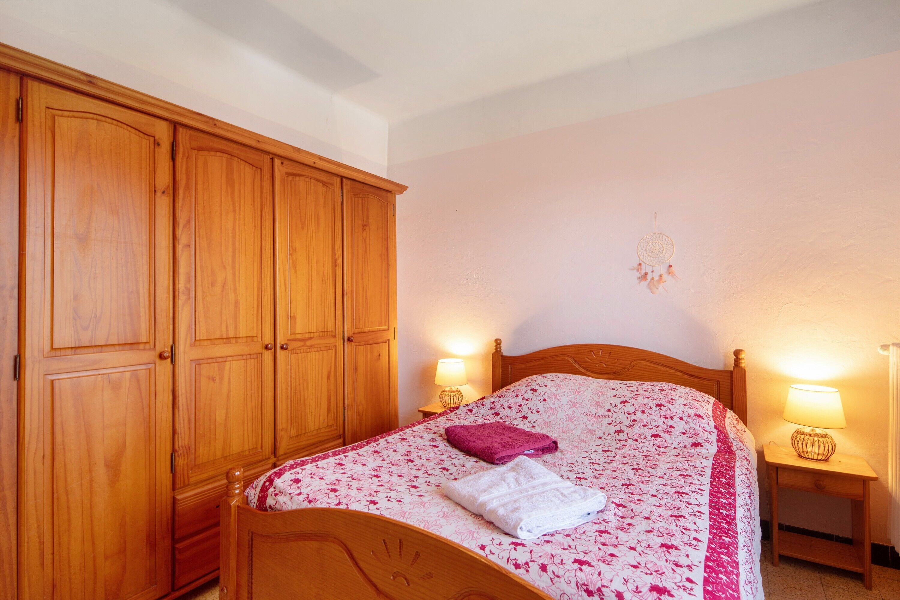 A bedroom with a wooden wardrobe, a bed with a floral bedspread, two bedside lamps, and a dreamcatcher on the wall.