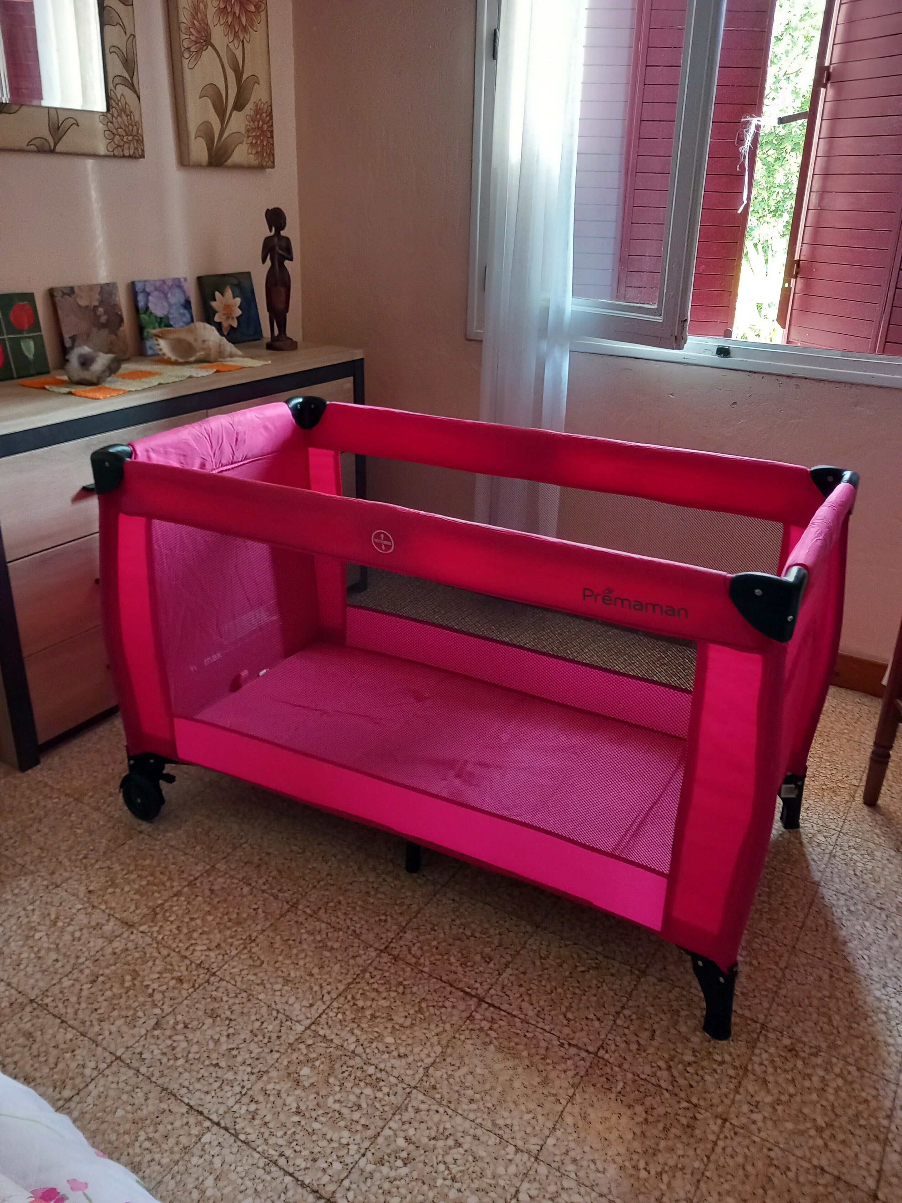 Un berceau rose dans une chambre avec une fenêtre, une commode et des décorations murales.