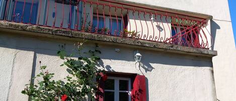 A building with a balcony, a small window with red shutters, and a flower box with red flowers.