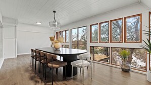 A spacious dining area with a round table and chairs, large windows with a view of trees, and a wooden ceiling.