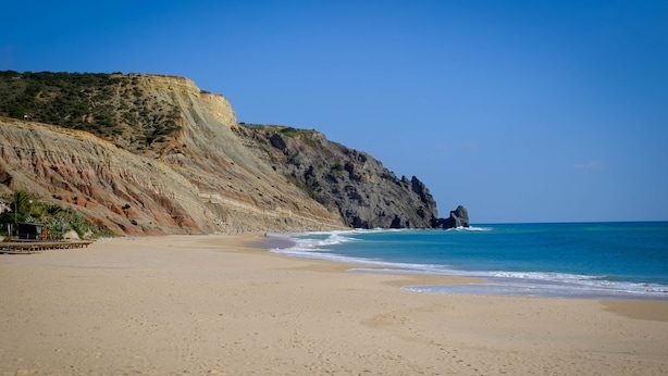 A beach with a rocky cliff, clear blue water, and a sandy shore.
