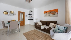 A living room with a brown sofa, a white armchair, a dining table with chairs, and a coffee table.